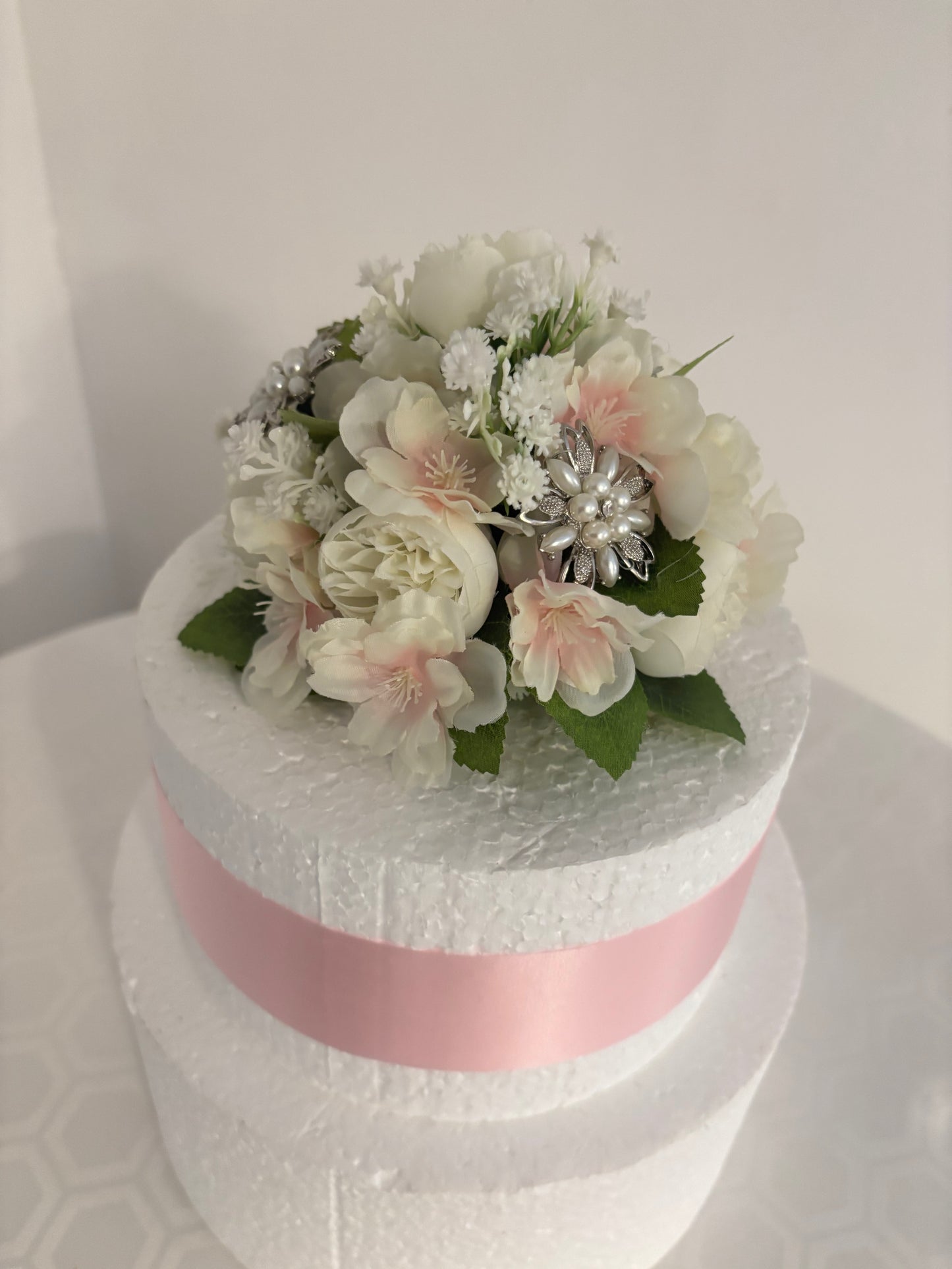 A two-tiered white artificial cake decorated with a pink ribbon and topped with a bouquet of white and pale pink artificial flowers with diamante & green leaves, set against a plain light background.