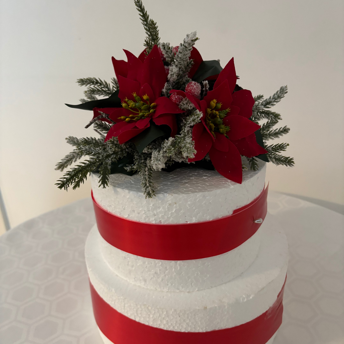A decorative round Christmas floral centre piece on top of  two tier white foam cylinder cake wrapped with red ribbons. The centre piece is hand made  with artificial red poinsettias and frosted greenery.