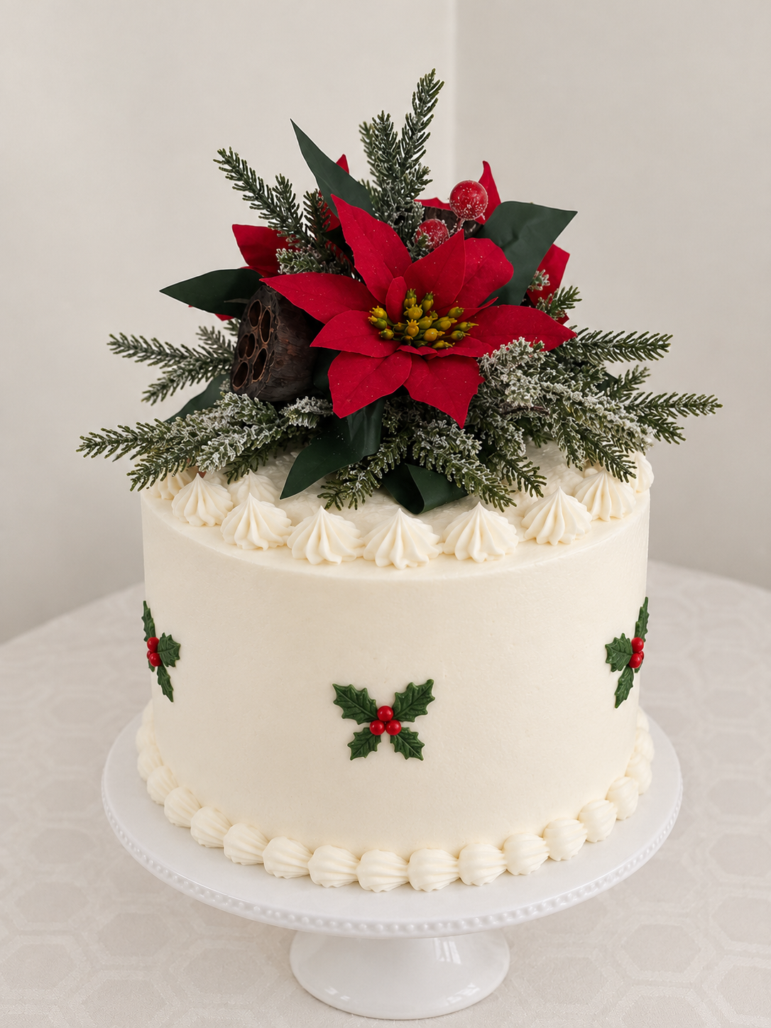 A round white frosted cake on a white pedestal stand, decorated with piped cream borders, holly motifs, and a large holiday cake topper featuring 3 red silk poinsettias, green leaves, artificial berries and snowy spruce branches.