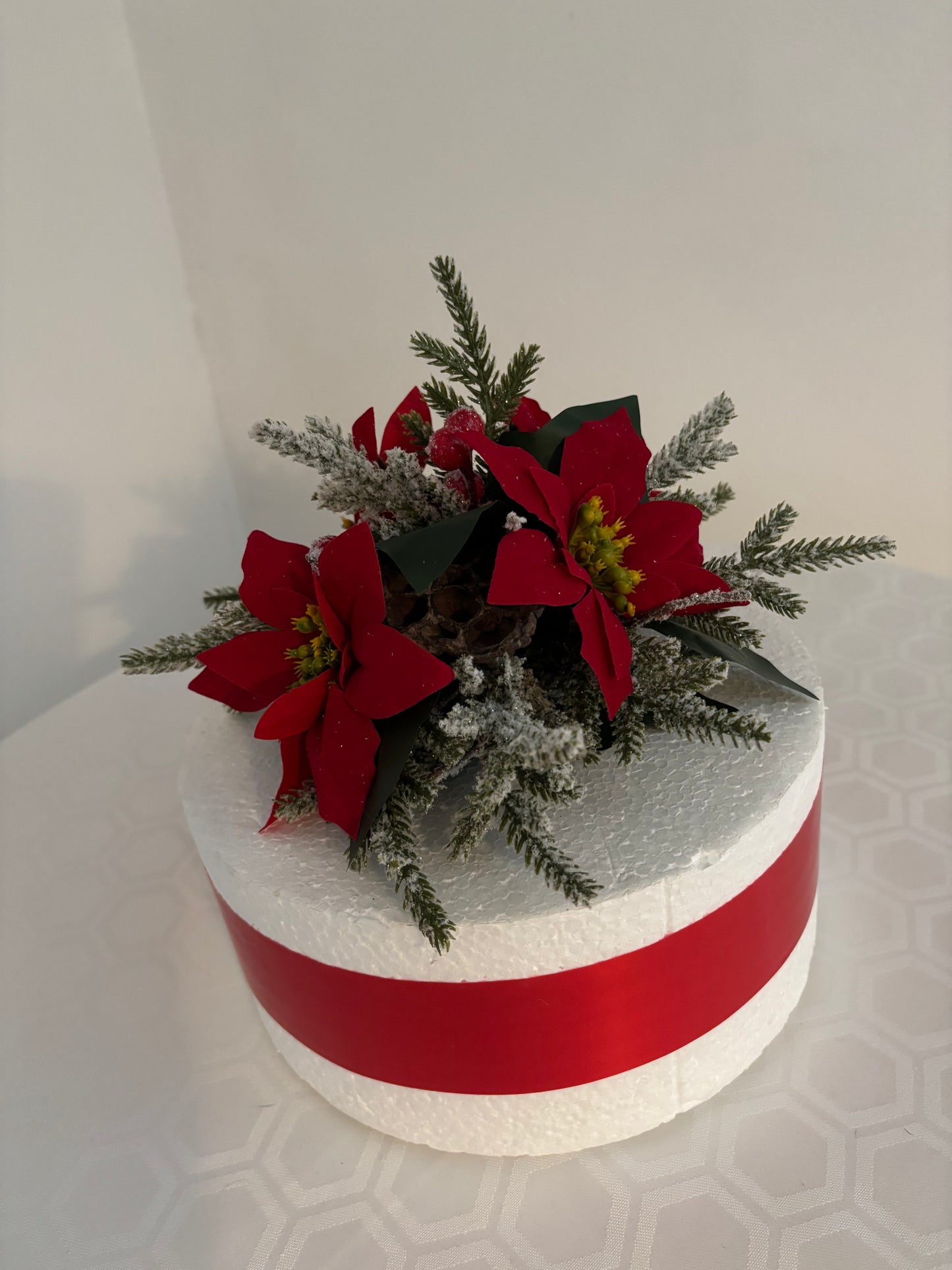 A round white cake base with a wide red ribbon band and a festive holiday floral decoration on top featuring red poinsettias, green spruce branches, and frosted greenery, shown against a plain light background.