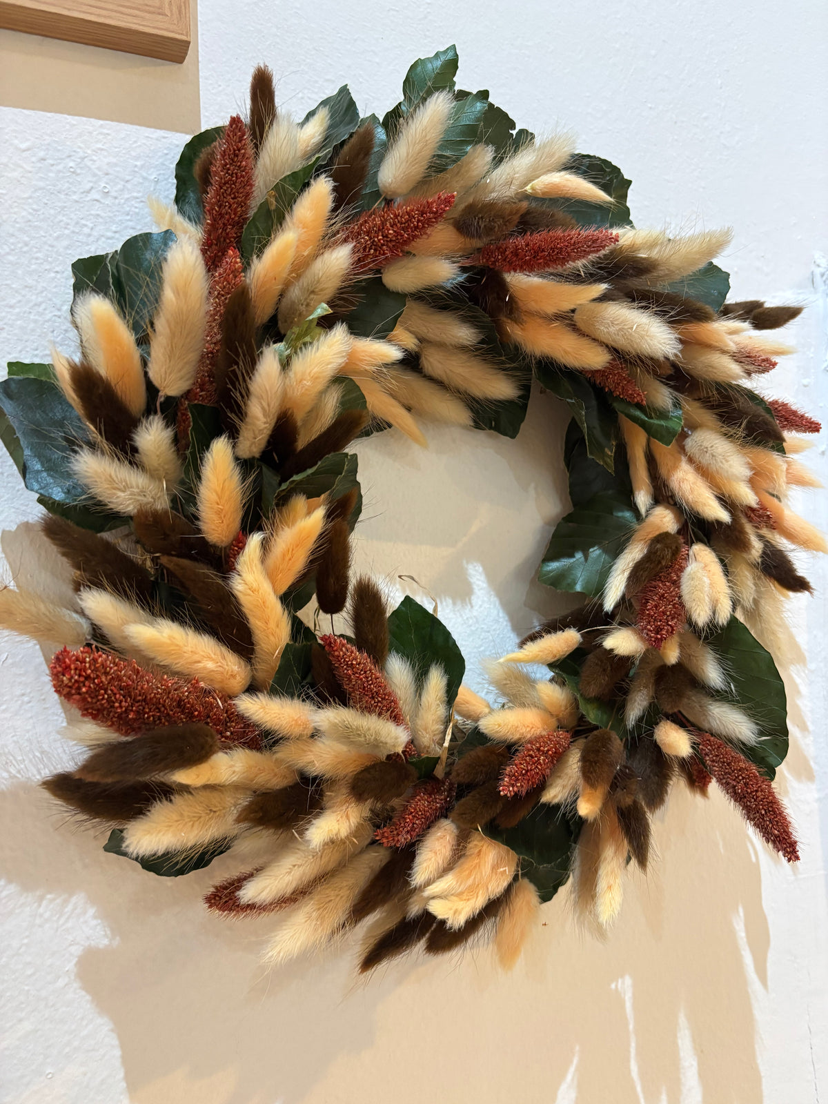 A decorative autumn wreath made of glossy green beech leaves, fluffy peach, beige and cream bunny tail grass, and rust-coloured millet pods, hanging on a white wall.