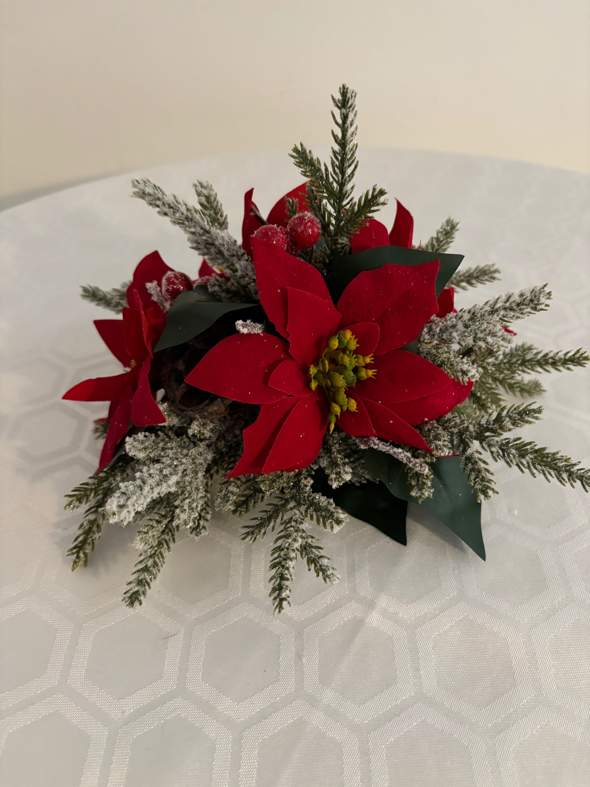A festive artificial floral centerpiece featuring bright red poinsettias, frosted green spruce branches, and small red berries.