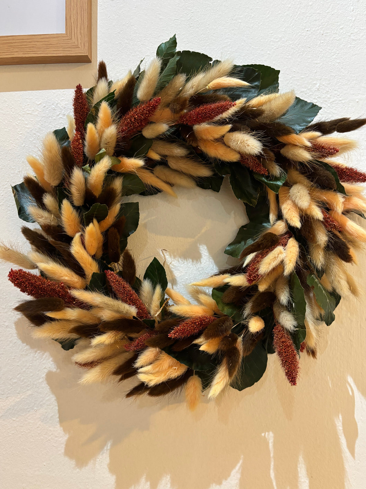 A decorative autumn wreath made of preserved green beech leaves, fluffy beige, peach and cream bunny tails, and rust-brown seed spikes, hanging on a warm off-white wall.