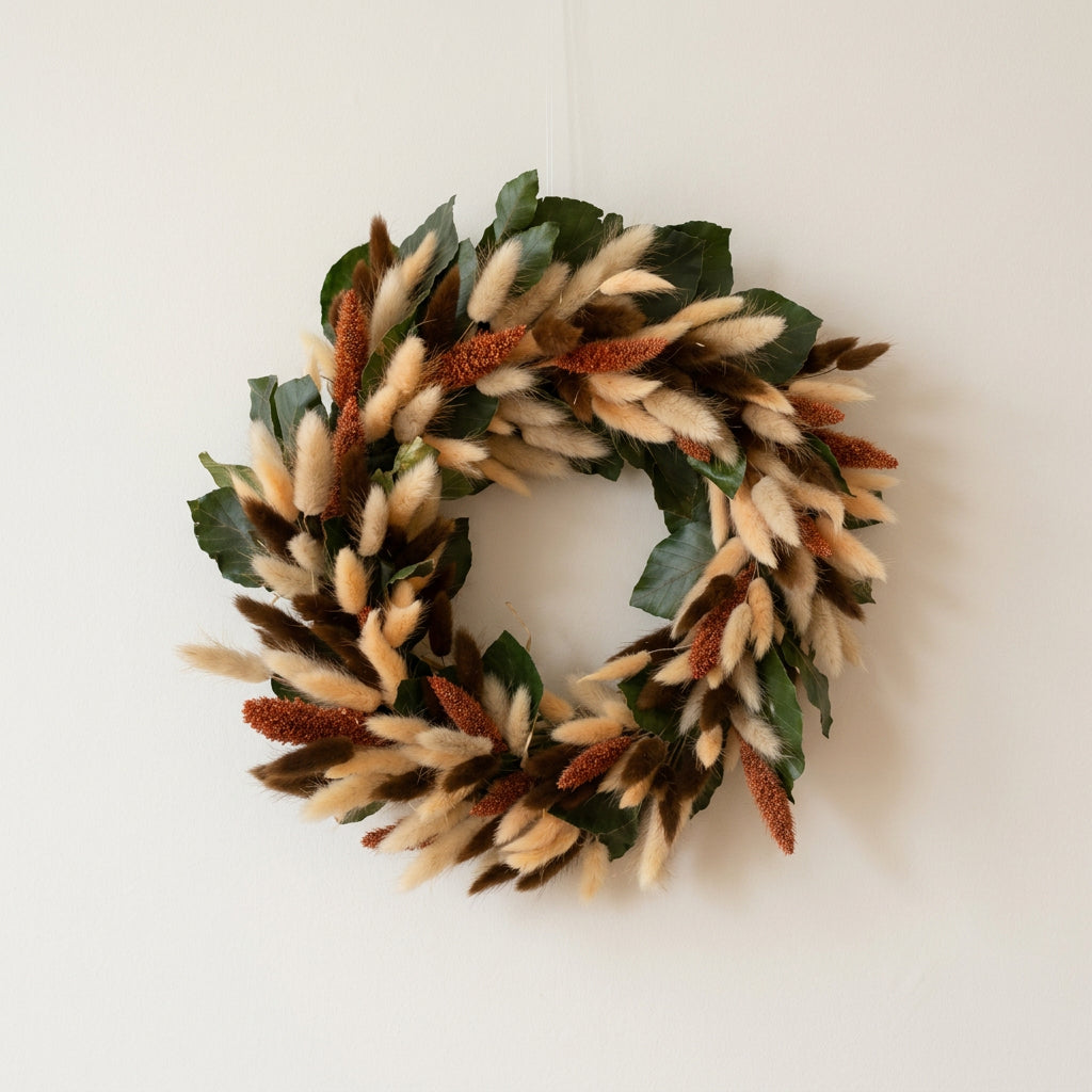 A decorative autumn wreath made of preserved green beech leaves, fluffy cream, peach and brown bunny tails, and rust-red decorative millet pods, hanging on a white wall.