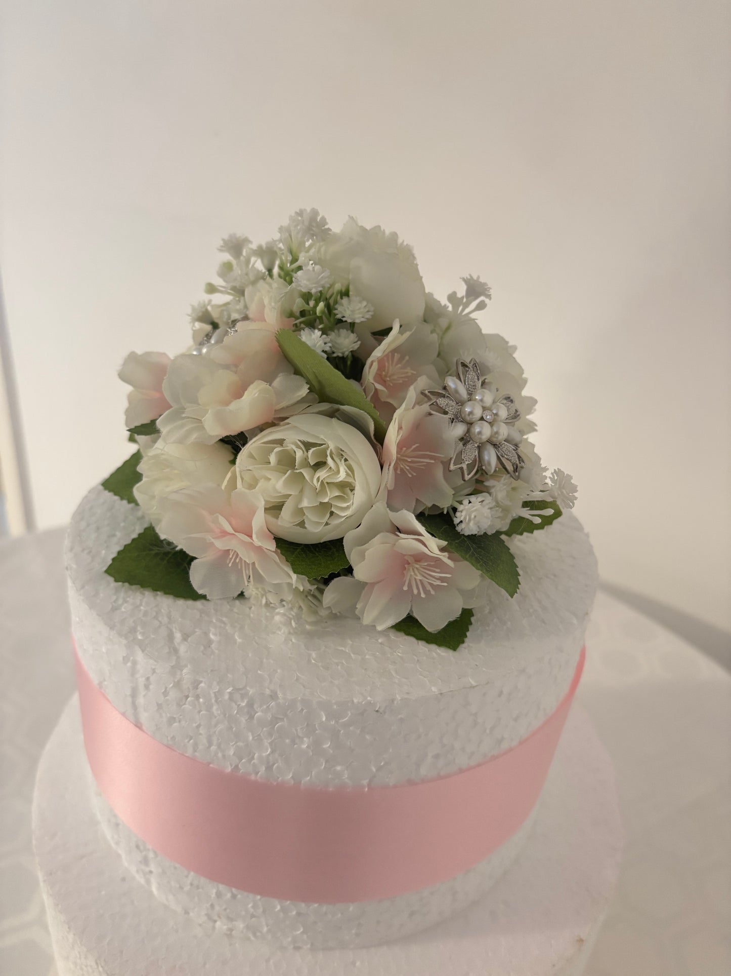 A round floral arrangement of ivory, white and pale pink flowers with green leaves, photographed against a plain light background.