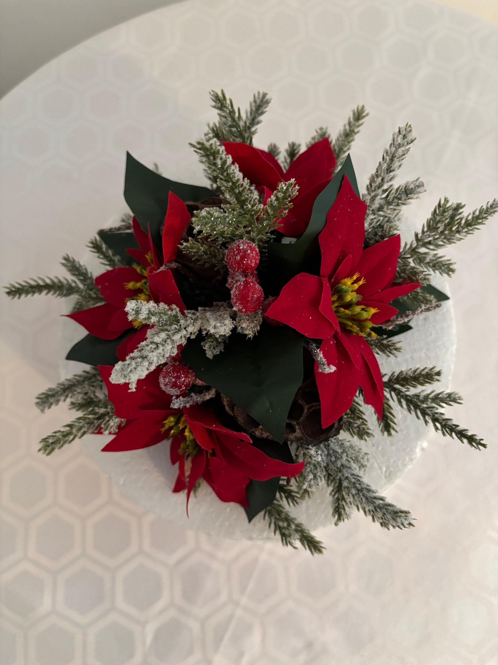 A festive artificial holiday floral arrangement featuring 3 red poinsettias,  green leaves, frosted spruce greenery, and red berries, photographed from above on a white patterned surface.