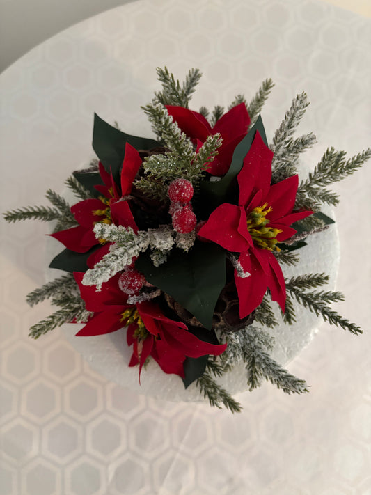 A festive artificial holiday floral arrangement featuring 3 red poinsettias,  green leaves, frosted spruce greenery, and red berries, photographed from above on a white patterned surface.