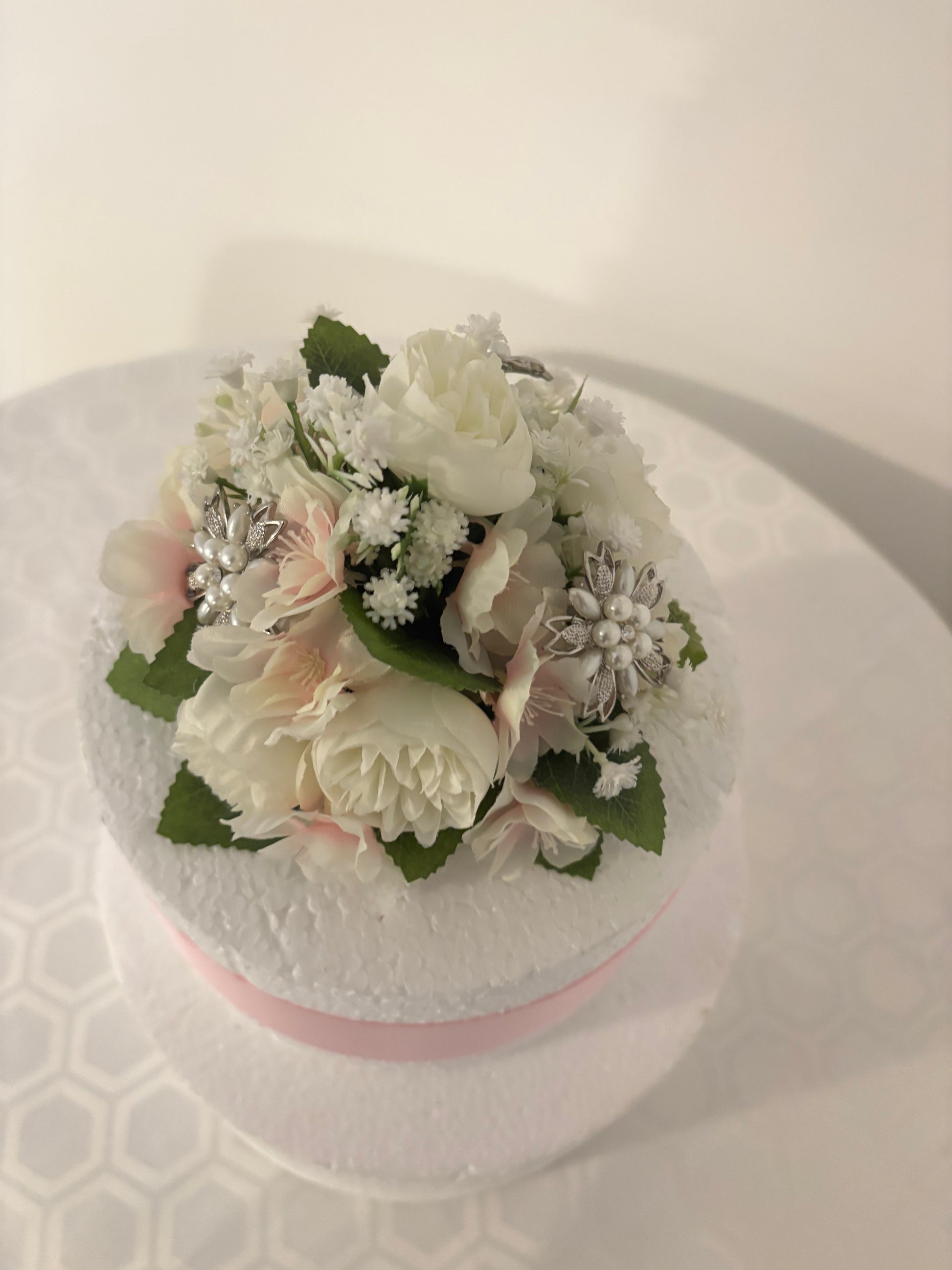 A round cake decorated with a bouquet of white and pale pink artificial flowers and green leaves, with pretty diamante decorations.
