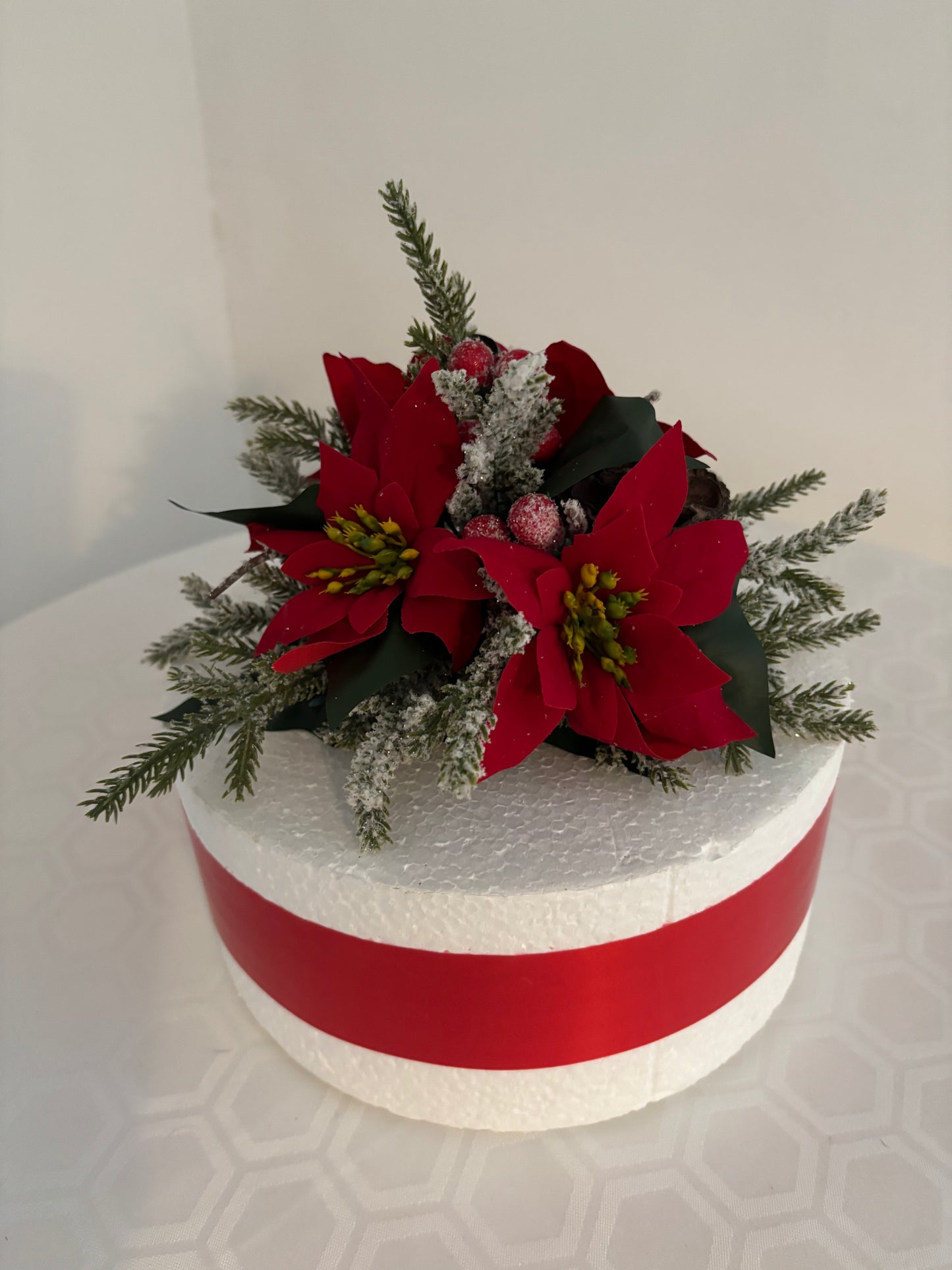 A round white cake with a cake topper decorated with red poinsettias, green pine branches, red berries, and a red ribbon band around the cake photographed against a plain light background.